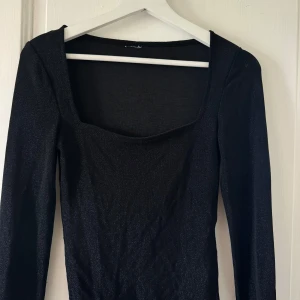 Svart glittrig långärmad topp - Snygg svart topp glittrigt tyg och lång ärm. Den har en fyrkantig urringning som ger en modern vibe och är figurnära i modellen. Perfekt för dig som vill sticka ut med lite extra shine. Passar dig som gillar stilrena plagg med en twist.