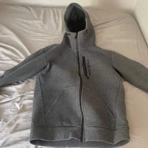 Sail racing zip hoodie - Säljer en grå hoodiejacka från Sailracing med hel dragkedja och praktisk bröstficka med zip. Jackan har en stor huva och är tillverkad i mjukt polyester-material, perfekt för chill eller utomhusaktiviteter. Snygg och enkel design med lång ärm och normal passform.