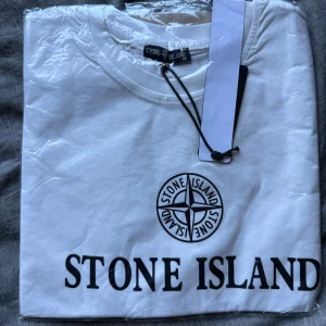 Vit Stone Island t-shirt med tryck - Vit t-shirt från Stone Island med klassiskt svart logotryck på bröstet och stor text under. Rund hals och korta ärmar, perfekt för en clean och stilren look. Materialet är mjuk bomull som känns skönt mot huden. En ikonisk tee för dig som gillar streetwear. Fortfarande kvar i påsen med tagg. Lite stor i storleken