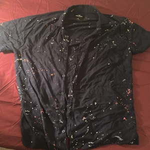 Svart och tunn Hollister skjorta med färgstänk - Svart skjorta från Hollister med korta ärmar och coolt färgstänksmönster.