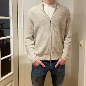 Beige stickad kofta från NN07 - Säljer en beige stickad kofta från NN07 med italienskt garn. Koftan har en snygg ribbad krage och dragkedja framtill. Perfekt för lager-på-lager och chill stil. Långärmad och med klassisk passform som funkar till allt.