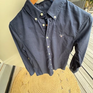 Mörkblå skjorta från GANT, 158/164 - Snygg mörkblå skjorta från GANT med klassisk button-down krage och broderad logga på bröstet. Skjortan har långa ärmar, vita knappar och är tillverkad i mjuk bomull. Perfekt för dig som gillar stilrena och tidlösa plagg. Jättefint Skick😁