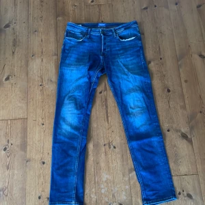 Blå slim straight jeans Jack & Jones - Snygga blå jeans från Jack & Jones i modellen Slim Straight. Klassisk femficksdesign med normal passform och raka ben. Jeansen har tydliga slitningar och kontrastsömmar, tillverkade i jeansmaterial. Perfekt för dig som gillar en stilren och enkel look. Defekt på sista bilden därmed priset. 