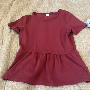 Vinröd peplum blus med volang - Supersöt vinröd blus med peplumvolang nedtill och rund halsringning. Toppen har korta ärmar och är gjord i mjuk bomull, perfekt för dig som gillar en chill och feminin stil. Volangen ger en extra touch och gör den enkel att styla med jeans eller kjol.