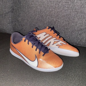 Nike Mercurial orange fotbollsskor - Säljer ett par Nike Mercurial fotbollsskor i orange med stora vita loggor på sidan och svart snörning. Skorna har en låg profil och platt vit sula med rosa detaljer undertill. Ovandelen är i syntetmaterial och designen är riktigt sportig och clean.
