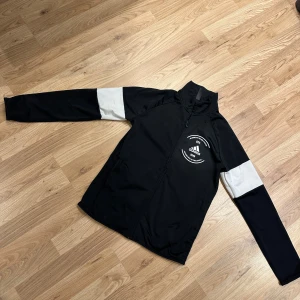Svart tröja från Adidas - Svart tröja från Adidas med vit logga och tryck på bröstet. Tröjan har hel dragkedja framtill och vita paneler på ärmarna som ger en sportig vibe. Tillverkad i polyester och har långärmad design med ståkrage. Perfekt för dig som gillar streetstyle och sport.