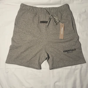 Grå Essentials shorts  - Gråa shorts från Essentials Fear of God med elastisk midja och snörning framtill. Mjukt bomullsmaterial, fickor på sidorna och en bakficka. Diskret logga tryckt på vänster ben. Perfekta för chill dagar eller träning.