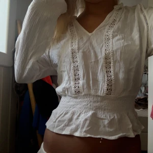 Vit blus med spets och volang - Säljer en superfin vit blus med v-ringning och långa ärmar. Blusen har snygga spetsdetaljer framtill och smockad midja som ger en cool volangkant. Materialet är lätt och krinklat, perfekt för dig som gillar romantisk stil med lite edge.