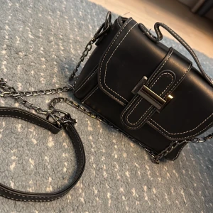 Svart axelväska med kedja och vita sömmar - Snygg svart axelväska med kedja och kontrasterande vita sömmar. Väskan har ett stilrent spänne framtill och flera fack inuti, inklusive dragkedja. Materialet är skinnimitation och den har både handtag och axelrem i metall och skinn.