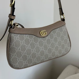Beige axelväska - Snygg beige axelväska med klassiskt GG-mönster och guldfärgad logodetalj framtill. Väskan har en justerbar axelrem i skinn och dragkedja upptill. 