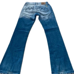 Blå bootcut jeans från Meltin'Pot - Snygga blå jeans från Meltin'Pot med bootcut passform och klassisk femficksdesign. Jeansen har ljusa slitningar och coola broderade detaljer på bakfickorna samt en brun logopatch. Perfekt för dig som gillar retrovibe och breda ben.