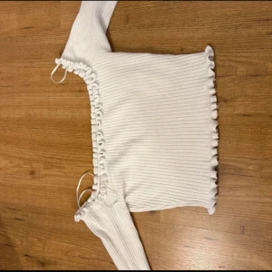 Vit ribbad topp från FB Sister - Supersöt off shoulder topp från FB Sister med långa ärmar och volangdetaljer längs kanterna. Toppen har en fyrkantig urringning och är croppad, vilket ger en trendig vibe. Perfekt för dig som gillar en feminin och stilren look.