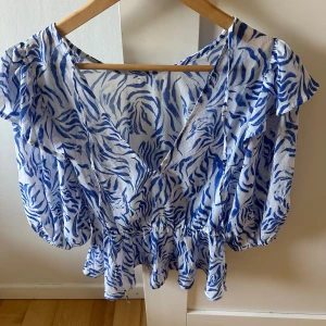 Blåvit blus S  - Säljer en blåvit blus v-ringad hals. Blusen har korta ärmar med volangdetaljer och ett elastiskt midjeparti som ger en snygg siluett. Materialet är lätt och luftigt, storlek S. Blusen är i nyskick 