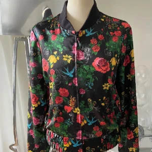 Blommig bomberjacka med dragkedja - Snygg bomberjacka med svart botten och färgglatt blommigt mönster i rött, grönt, gult och blått. Jackan har ribbad krage och muddar samt dragkedja framtill. Materialet är glansigt och känns lätt mot kroppen, perfekt för dig som vill sticka ut med en cool look.