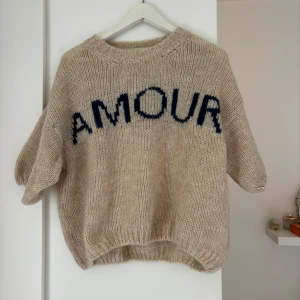 Beige stickad tröja med AMOUR - Säljer en oversized stickad tröja från Design by Si i beige med mörkblå texten 'AMOUR' framtill. Tröjan har rund hals, ribbade muddar och kortare, lite puffiga ärmar. Perfekt för dig som gillar chill och trendiga statement-plagg.
