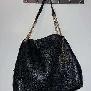  axelväska Michael Kors  - Snygg svart axelväska från Michael Kors i äkta skinn med guldfärgade kedjor och MK-hänge. Väskan har en mjuk, rundad form och detaljer i guld. Perfekt storlek för allt du behöver och med dragkedja på insidan.
