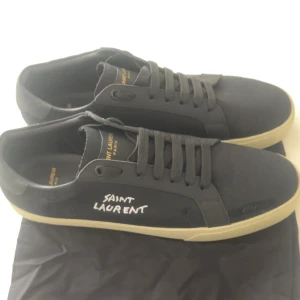 Svarta Saint Laurent sneakers strl 45 - Svarta sneakers från Saint Laurent med vit logga på sidan och guldtext på plösen. Snygg kontrast med beige sula och klassisk snörning. Ovandelen är i textil och skinn, och sulan har fiskbensmönster. Perfekt för dig som gillar stilrena och exklusiva sneakers.