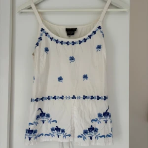 Vit blommig linnetopp från Passport - Supersöt vit linnetopp med blå broderade blommor och smala axelband från Passport. Toppen har knytband i ryggen och detaljerade blommor längs nederkant och halsringning. Perfekt för varma dagar och ger en fräsch vibe till din outfit.