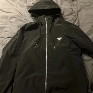 Svart vindjacka från Prada - Svart vindjacka från Prada med huva och silverfärgad dragkedja. Jackan har en clean look med ikonisk Prada-logga på bröstet och justerbara snören vid huvan. Materialet är lätt och syntetiskt, perfekt för dig som gillar stilren design och exklusiva märken.