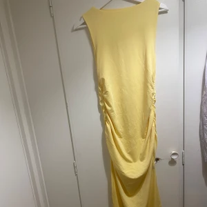 Gul långklänning med drapering - Snygg gul långklänning med ärmlös design och draperade detaljer längs sidorna. Klänningen har en slits nedtill för extra rörelsefrihet och är gjord i ett mjukt, lätt material som känns skönt mot huden. Perfekt för dig som vill sticka ut med en färgglad och stilren look. Aldrig använd. Storlek S/M