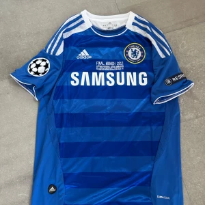 Chelsea FC Drogba 2012 matchtröja - Chelsea FC fotbollströja från Champions League-finalen 2012 med Drogba och nummer 11 på ryggen. Blå med vita detaljer, Adidas-logga, klubbmärke och Samsung som sponsor. Kortärmad med randigt mönster och CL-patch på ärmen. Tillverkad i lätt och ventilerande polyester.helt ny med tagg en replika 