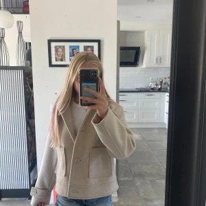 Beige kavajjacka från 157 - Snygg beige kavajjacka från 157 med klassisk krage och två stora fickor framtill. Jackan har knappar både fram och vid ärmsluten, och är tillverkad i mjukt ullmaterial. Perfekt för dig som gillar stilrena och enkla plagg med lite oversized känsla.