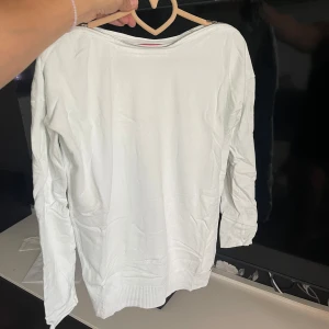 Vit långärmad tröja från Guess - Vit långärmad tröja från Guess med rund hals och snygg dragkedjedetalj längs axlarna. Tröjan är i mjuk bomull och har ribbade muddar vid ärmslut och nederkant. Perfekt för dig som gillar stilrena och enkla plagg med en twist.