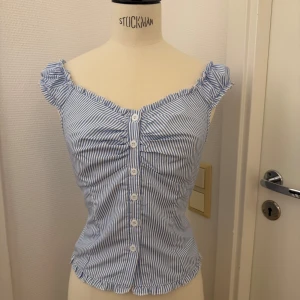Blåvit randig topp - Supersöt topp med blåvita ränder och knappar framtill. XS. Toppen har volangdetaljer vid axlar och nederkant, samt en lätt rynkad effekt över bysten. Kan hämtas på Hägerstensåsen om du vill slippa frakt. 🌸