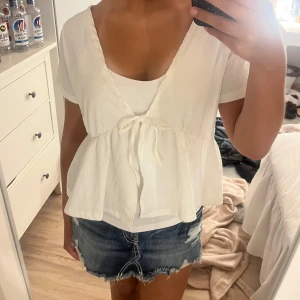 Vit blus med knyt och v-ringning - Supersöt vit blus med v-ringning och knyt framtill. Blusen har korta ärmar och en luftig, flowy passform som ger en avslappnad vibe. Perfekt till jeansshorts eller kjol för en fräsch look. Materialet känns lätt och svalt, perfekt för varmare dagar.