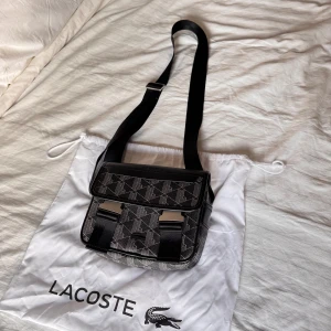 Svart axelväska från Lacoste - Snygg svart axelväska från Lacoste med geometriskt mönster i vitt. Väskan har justerbar rem, två metallspännen framtill och dragkedja. Insidan har en orange detalj och flera fack för dina prylar. Perfekt för dig som vill ha en stilren och praktisk väska.