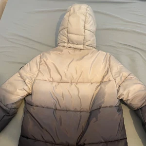 Grå pufferjacka från H&M med huva - Snygg pufferjacka från H&M i en gradient från ljusgrå upptill till mörkgrå nedtill. Jackan har en stor, vadderad huva och dragkedja framtill. Fickor på sidorna och quiltad design ger en modern look. Perfekt för kalla dagar och enkel att matcha med streetwear.