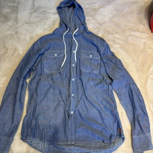 Blå Fishbone skjorta med huva - Snygg blå skjorta från Fishbone med huva och vit snörning. Skjortan har två bröstfickor med knappar och ett rutigt foder i huvan och nederkant. Långärmad och tillverkad i mjuk bomull, perfekt för en chill och avslappnad stil.