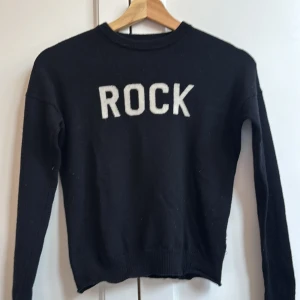 Svart kashmirtröja från Zadig & Voltaire - Cool svart stickad tröja från Zadig & Voltaire med vit text 'ROCK' framtill. Tröjan har rund hals, ribbade muddar och är långärmad. Tillverkad i mjuk kashmir som känns lyxig och skön mot huden. Perfekt för dig som gillar statement-plagg och vill sticka ut.