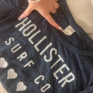 Mörkblå Hollister t-shirt med broderi - Snygg mörkblå t-shirt från Hollister med stora broderade bokstäver och hjärtan i vitt på framsidan.  Finns lite skavanker då trådar har ramlat upp.  Klassisk rund hals och korta ärmar. Mjuk bomullskvalitet som är skön mot huden. Perfekt för dig som gillar surf vibes och tydliga loggor.