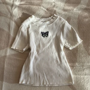 Vit ribbad topp med fjärilsbroderi - Vit ribbad topp från H&M med korta ärmar och rund hals. På bröstet sitter ett broderat fjärilsmotiv i svart och lila toner som ger en söt detalj. Materialet är mjukt och stretchigt, perfekt för en chill och stilren look. Passformen är normal och toppen är enkel att matcha med jeans eller kjol.