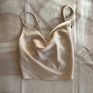 Satin korsettopp från Gina Tricot - Snygg beige satin korsettopp från Gina Tricot med smala axelband och v-ringad front. Toppen har snörning i ryggen för en justerbar passform och är croppad. Materialet är mjukt och har en lätt glansig yta som ger en fräsch vibe.