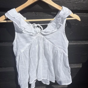 Vit volangblus i linne med knyt Gina Tricot young stl.146 - Gina Tricot Young stl.146. Supersöt vit blus i linne med volangdetaljer längs axlarna och ett romantiskt knyt i nacken. Luftig och flowy passform, perfekt för varma dagar. Ärmlös modell med lätt rynkade partier som ger en bohemisk vibe.