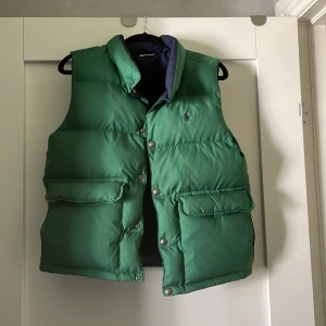 Grön och marinblå dunväst från Polo Ralph Lauren - Säljer en fet dunväst från Polo Ralph Lauren, vändbar med grön utsida och marinblå insida. Ena sidan har Polo-loggan broderad på bröstet, andra sidan har en stor vit R-applikation. Västen har tryckknappar, hög krage och stora fickor framtill. Perfekt för lager-på-lager under höst och vinter.