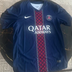 PSG matchtröja D.Doué #14 Nike - Paris Saint-Germain fotbollströja med D.Doué och nummer 14 på ryggen. Marinblå med röd och vit randigt geometriskt mönster i mitten, PSG-logga på bröstet och Nike Swoosh. Qatar Airways tryck på framsidan. Tillverkad i lätt polyester med korta ärmar och V-ringad hals.