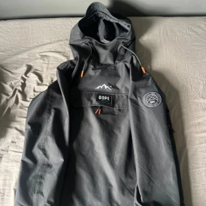 Dope jacka - Svart vindjacka från Dope Snow med huva och coola orangea detaljer på snörningen. Stor ficka framtill med logotyp och snyggt märke på ärmen. Perfekt för dig som gillar streetstyle och outdoor-vibe. Jackan är i polyester och har en loose passform.