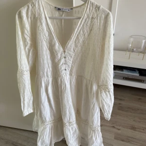 Vit kortklänning med spets från Zara - Supersöt vit kortklänning från Zara med v-ringning, spetsdetaljer och knappar framtill. Klänningen har långärmade ärmar med spetsinslag och ett diskret prickigt mönster i tyget. Perfekt för dig som gillar romantisk och boho-inspirerad stil.