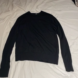 Svart stickad tröja från Uniqlo - Svart stickad tröja från Uniqlo i storlek S, tillverkad av 100% ull. Tröjan har en klassisk rund hals och långa ärmar med ribbade muddar vid ärmslut och nederkant. Perfekt för dig som gillar enkel och stilren design.