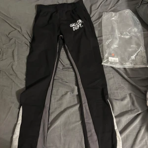 Svarta Gallery Dept. mjukisbyxor - Svarta mjukisbyxor från Gallery Dept. med vit logga på framsidan och gråa samt ljusgrå sidopaneler längs benen. Byxorna har elastisk midja och är gjorda i 100% bomull, vilket gör dem riktigt sköna och avslappnade. Perfekta för chill eller streetwear.