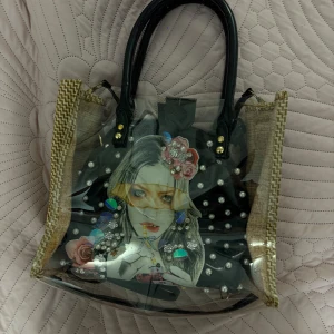 Transparent handväska med pärlor och tryck - Säljer en unik transparent handväska med svarta handtag och beige kantdetaljer. Inuti finns en mindre svart väska med coolt tryck av en tjej, dekorerad med pärlor, blommor och glittriga stenar. Perfekt för dig som gillar att sticka ut med din stil.