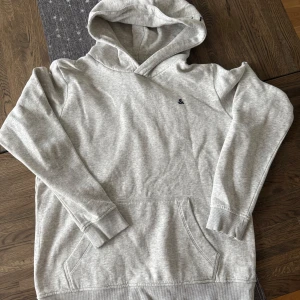 Grå hoodie från Jack & Jones - Säljer en stilren grå hoodie från Jack & Jones. Den har en klassisk känguruficka och en bekväm huva med logotyptryck. Perfekt för en avslappnad stil. Storlek 176
