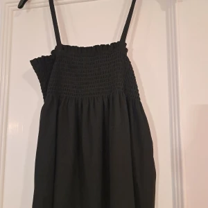 Svart topp från Ellos - Säljer en stilren svart topp från Ellos med smockdetaljer upptill och smala axelband. Perfekt för en avslappnad look. Passar bra till både vardag och festliga tillfällen.