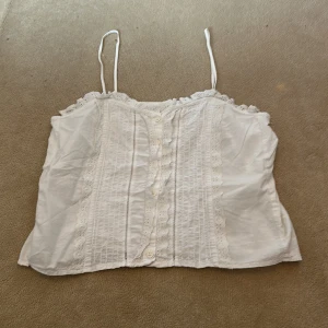 Vit linne med spetsdetaljer - Säljer en söt linne med smala axelband och spetsdetaljer längs kanterna. Toppen har knappar framtill. Har aldrig använt den. 