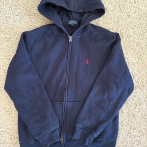 Mörkblå hoodie från Polo Ralph Lauren - Säljer en mörkblå hoodie från Polo Ralph Lauren med dragkedja och huva. Den är i väldigt bra skick och passar bra nu till våren/sommaren