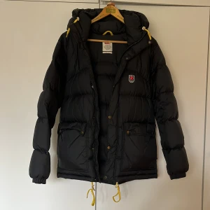 Fjällräven expedition down jacket - Säljer en svart dunjacka från Fjällräven med huva och gula detaljer. Jackan har dragkedja och knappar framtill samt ribbade muddar vid ärmsluten. Använd en säsong, pack påsen som jackan kom med ingår. 