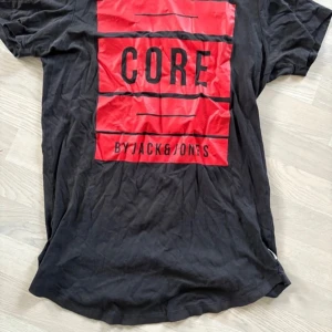 Svart t-shirt från Jack & Jones - Säljer en svart t-shirt från Jack & Jones med ett stort rött tryck på framsidan där det står 'CORE'. Perfekt för en avslappnad stil. T-shirten har korta ärmar och en rund halsringning. 